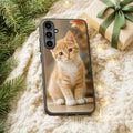 Capa de Samsung com a foto de um gatinho ruivo e branco, sobre uma manta creme com pinheiro