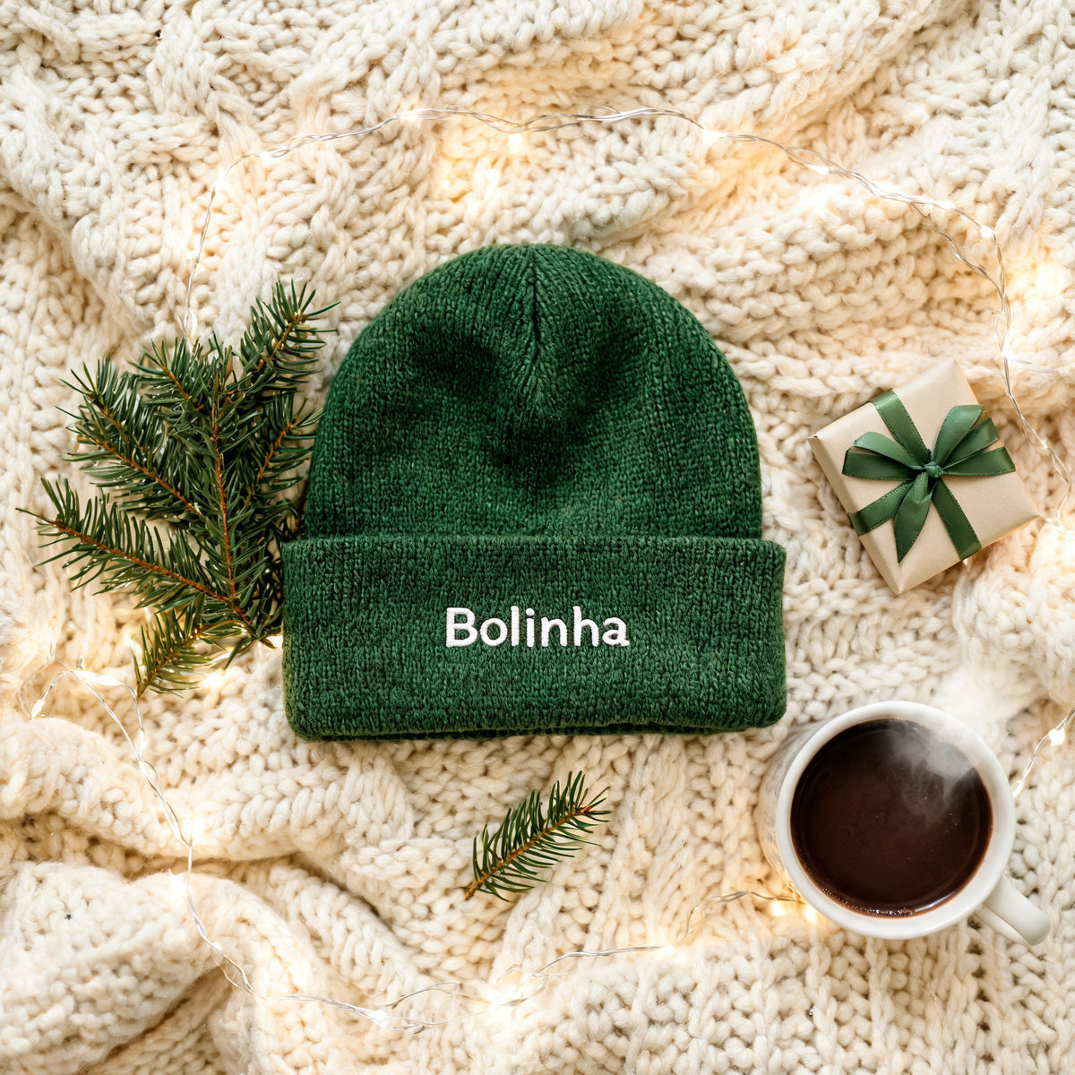 Gorro verde-escuro com o nome Bolinha bordado, sobre uma manta de malha creme com ramos de pinheiro
